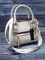 Front and side view of the white Christian Siriano purse showing two handles, adjustable strap, front zip pocket, and gold logo plate.