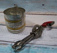 Photo of the vintage Foley Sift Chine cylindrical metal flour sifter with yellow horizontal stripe and branded text.