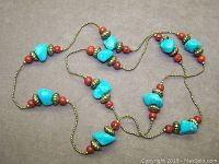 Photo showing the full length of the necklace laid out in a loose pattern on a neutral background. Necklace features large bright blue turquoise howlite stone beads, small smooth red accent beads, and antique brass-colored spacer beads on a brass bead chain.