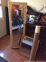 White framed mirror standing vertically to the left, gold framed mirror smaller leaning in front, reflective surfaces visible with some room elements reflected.