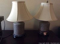 Pair of Asian style table lamps with beige fabric shades on a dark wooden surface against wall, one missing finial.
