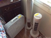 Three fans grouped together near a window: one square Lasko box fan and two vertical tower fans, one shorter and one Honeywell taller model.