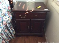 Full view of the wood side table cabinet nightstand showing the top surface, single drawer with brass-style handle, and two-door lower cabinet. Light reflections across the top surface.