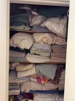 Three shelves filled with folded towels, linens, and queen-size sheets stored in a wall cubby, showing various colors and fabrics tightly stacked.