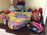 Image shows a variety of pet items arranged on a wooden surface, including large sealed bags of Hartz Home Protection piddle pads, small bags of Pup-Peroni mini dog treats, pink bandanas, dog toys, and grooming products in bottles and tins. An American flag is in the background.