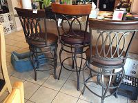 Three metal and wood swivel stools in a kitchen. One taller bar height stool and two shorter counter height stools. Brown padded seats and wooden top rails with metal circular back designs.