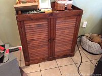 Front view of the vintage wood cabinet with two shutter style doors closed, showing design and size relative to tiles on floor.