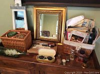 Display of vintage gold framed mirror on wooden dresser with wicker baskets containing books, perfumes, sunglasses, and small collectibles