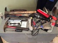 Photo showing various hand tools including hammers, putty scraper, screwdriver, paintbrush, pick hammer, and a Dremel Multi-Max rotary tool with power cord arranged on a gray wooden surface.