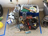 Photo showing assortment of tools and containers arranged on carpeted floor, includes hand tools, tool bag, plastic tub, and boxed items