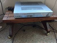 Photo showing vintage wooden slatted bench with Norcent DVD amplifier placed on top, carpeted floor background.