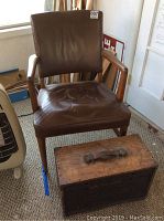 Wooden chair with armrests upholstered in brown leather showing signs of heavy wear placed behind wooden tool chest with leather handle on top.