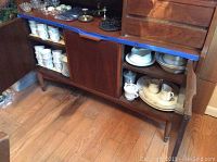Wide view of sideboard cabinet displaying multiple stacks of porcelain china including cups, plates, and bowls arranged on shelves.