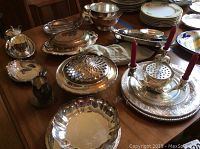Photo showing entire collection of silver plated serveware on wooden table, including covered dishes, platters, sugar and creamer set, candelabra with red candles and other serving pieces.
