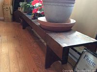 Side angle showing the entire bench with plants placed on top, hardwood floor beneath, showing its length and construction.