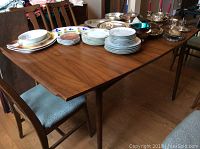 View of the entire rectangular dining table showing wood surface with stacked dishes and silverware on top, revealing general size and condition.