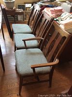 Six Danish Modern Mid Century Modern wooden dining chairs arranged in a row showing one wooden armchair with five armless chairs with light green patterned fabric upholstery, placed on a wooden floor with surrounding furniture and textiles.