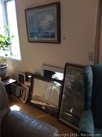 Room corner with multiple framed prints and mirrors stacked against the wall and a large framed print hanging above near a window