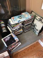 Pile of CDs and DVDs stacked on a bench near a fireplace, with vinyl record covers visible behind showing Eric Clapton and other album covers.