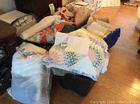 View of four large plastic bins with lids and one bag filled with mixed linens, towels, and clothing, including folded quilts and other fabric items.