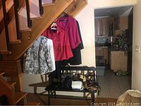 Photo of three women's coats hanging under staircase and five purses on a bench below them, showing the lot contents including various outerwear and handbags.