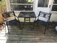 Photo showing black metal square mesh table with umbrella hole and two matching chairs with decorative backs arranged on wooden porch.
