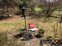 Wide view of outdoor items including bird feeder, metal chair with red pillow, two planters, cast iron decorative wheel on the ground, in an outdoor garden setting.