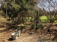 Photo of outdoor garden area showing black metal arbor, plastic planters, buckets, bird bath, and hanging objects