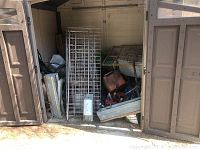 Storage shed interior showing stacked wire grid shelving panels leaning, red gas can on right, assorted tools and containers underneath
