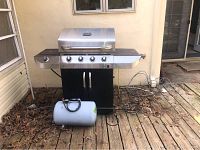 Front view of Char Broil Commercial Series grill with four control knobs, black cabinet base, side shelves, and white external propane tank in front on wooden deck.