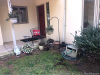 Front porch scene with black metal chair and side table, ceramic and plastic planters, garden hose caddy and shepherd hooks.