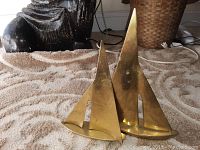 Two brass sailboat sculptures on a patterned carpet with various background objects. The taller sailboat has three sails, the shorter has two.