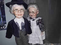 Front view of two porcelain dolls representing an elderly couple, detailed facial features, clothing, and accessories.