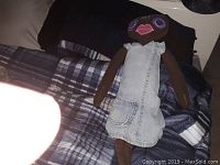Full view of the homemade fabric doll seated on a patterned bedspread, showing dark brown fabric body, denim dress, large embroidered purple eyes and large pink lips.