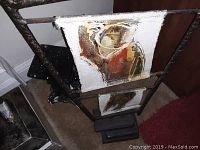 Two close-up photos of folk art paintings on canvas mounted on a rustic metal stand. The paintings depict abstract human forms with muted earth tones.