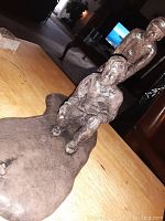 Side view of wood plated plague statue on wood tabletop, showing seated and standing figures with a textured, rough finish.