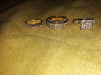 Three gold-colored rings on fabric background showing slim wedding band, wider band with two rows of stones, and a ring with rectangular cluster of stones.