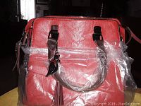 Front view of red handbag showing textured finish, handles wrapped in plastic, tassel and charm detail.