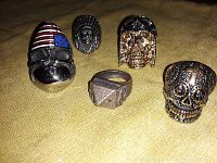 Five metal rings placed on a surface, including three featuring skull designs: one with an American flag helmet, a gold-tone skull with goggles, and a detailed black skull ring.