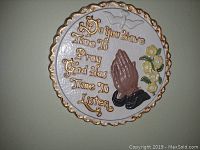 Photo of circular wall prayer plate with gold scalloped edges, 3D praying hands, flowers, and gold text on white background.