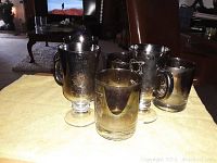 Photo showing six vintage silver-toned glasses with handles and pedestal bases set on a table, exhibiting tarnish and wear.