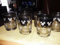 Six clear glass tumblers with silver Mickey Mouse head silhouettes on a light surface, arranged in two rows with three glasses in front and three behind.
