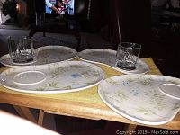 Photo shows four ceramic plates with floral design and two clear glass cups set on a wooden table with yellow cloth.