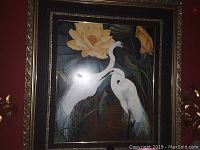 Framed art print showing two white herons with yellow flowers and green foliage. The ornate frame is dark with gold detailing around the edges.