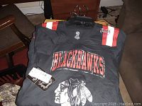 Folded black Blackhawks team shirt with red and white stripes on shoulders and Blackhawks logo, lying on a couch.
