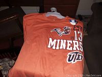 Orange UTEP Miners T-shirt on hanger laid flat showing front design with EST 1913, miner logo, and Miners UTEP text