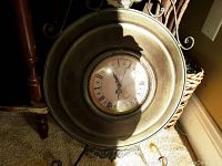 Antique round metal wall clock with Roman numeral dial, shown with sunlight and shadows highlighting the metal finish.