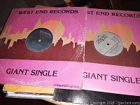 Two West End Records giant single vinyl records in pink and brown sleeves with a city skyline graphic. Black and gray center labels visible on vinyl.