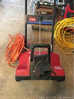 Front view of Toro Power Curve snow thrower showing red body, black chute, and orange power cord coiled beside it.