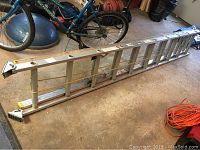 Full view of the metal extension ladder laid on the ground with bike, basketball and extension cord nearby.